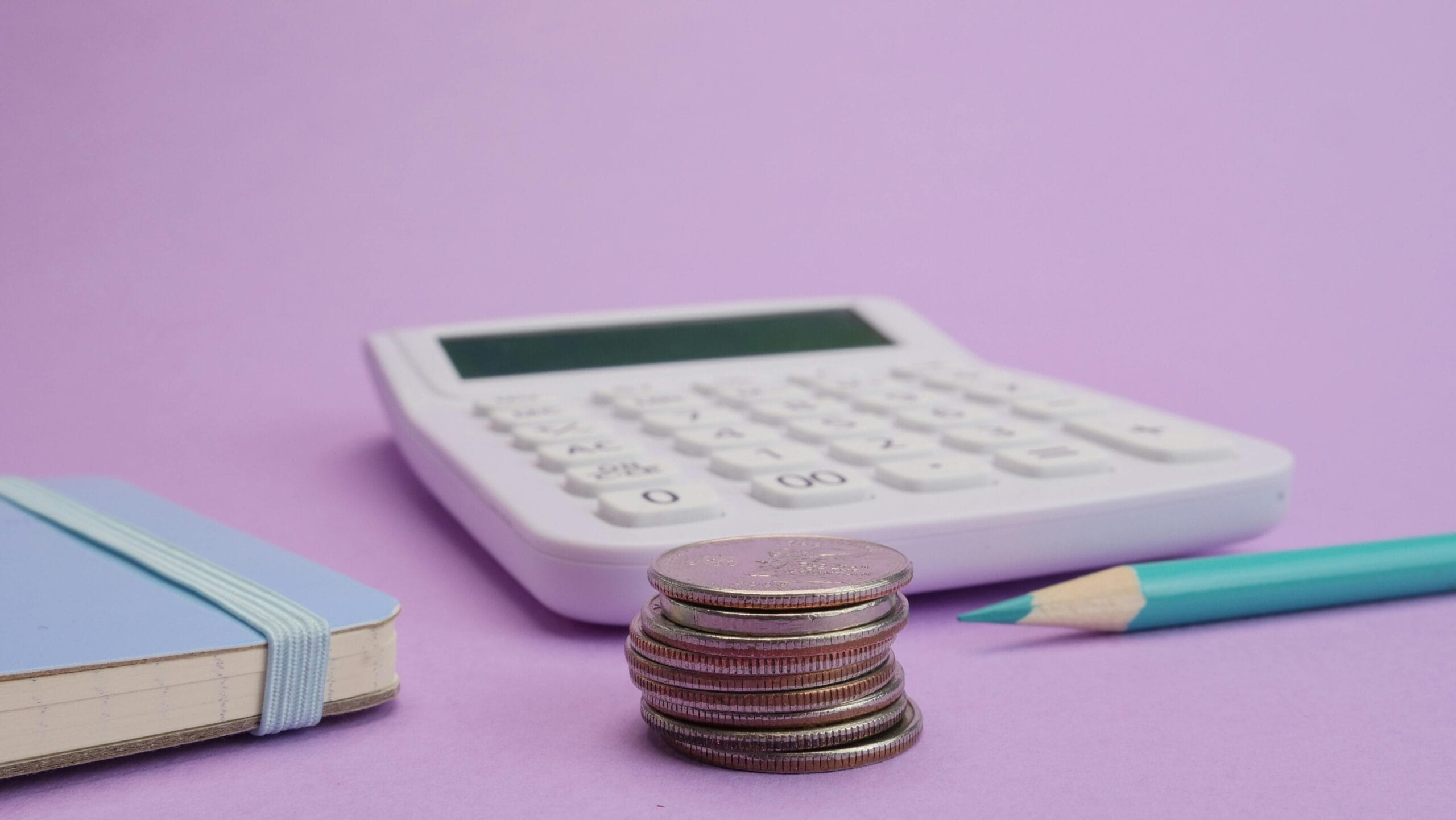 A stack of coins next to a calculator and a notebook with a pencil on a purple background.