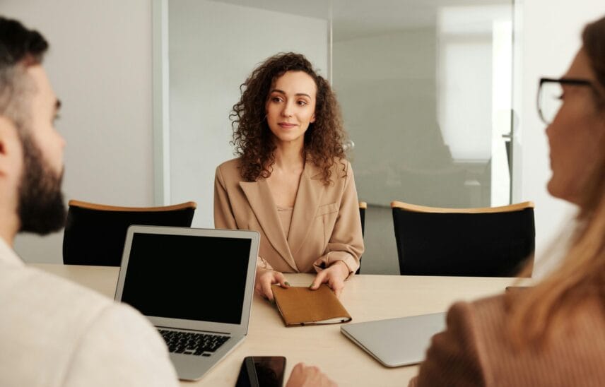 A woman with curly hair sitting at a table during a business meeting, holding a notebook.