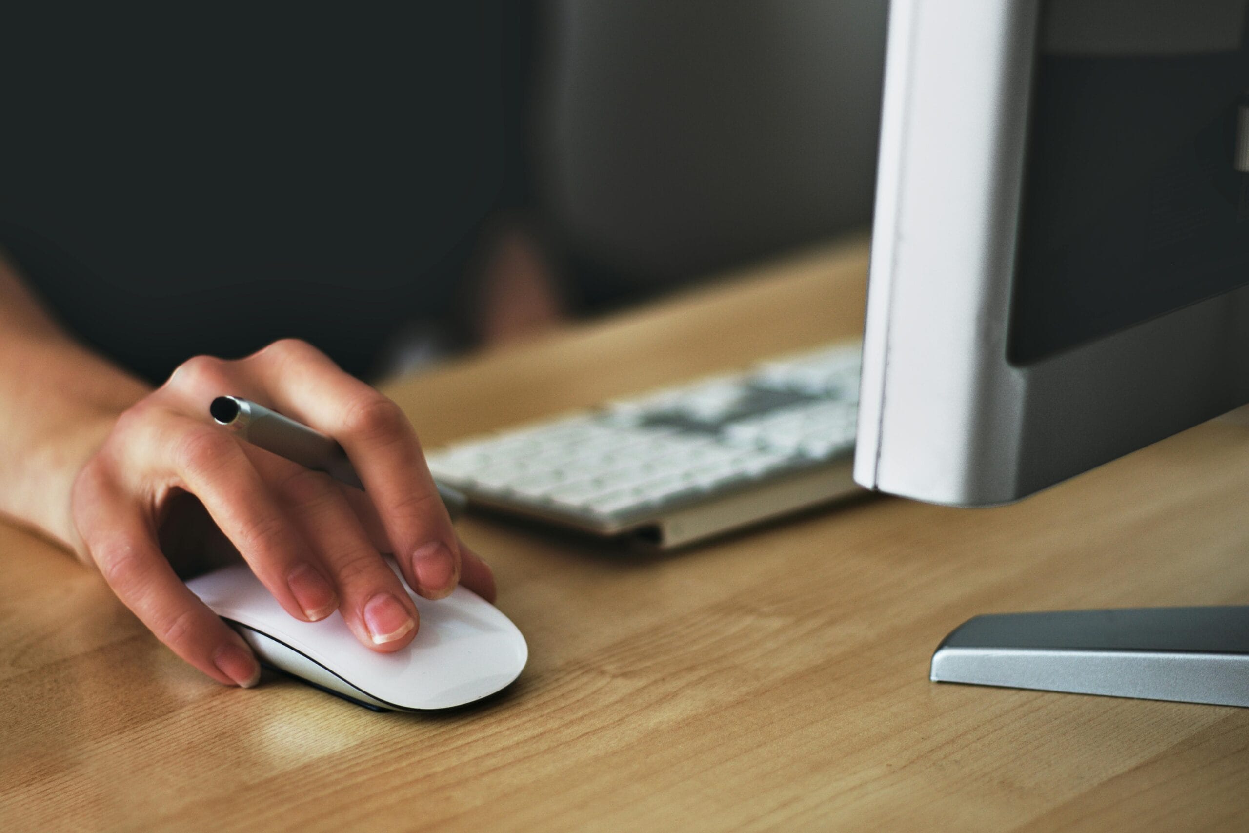 A hand holding a computer mouse while a keyboard and monitor are visible in the background.