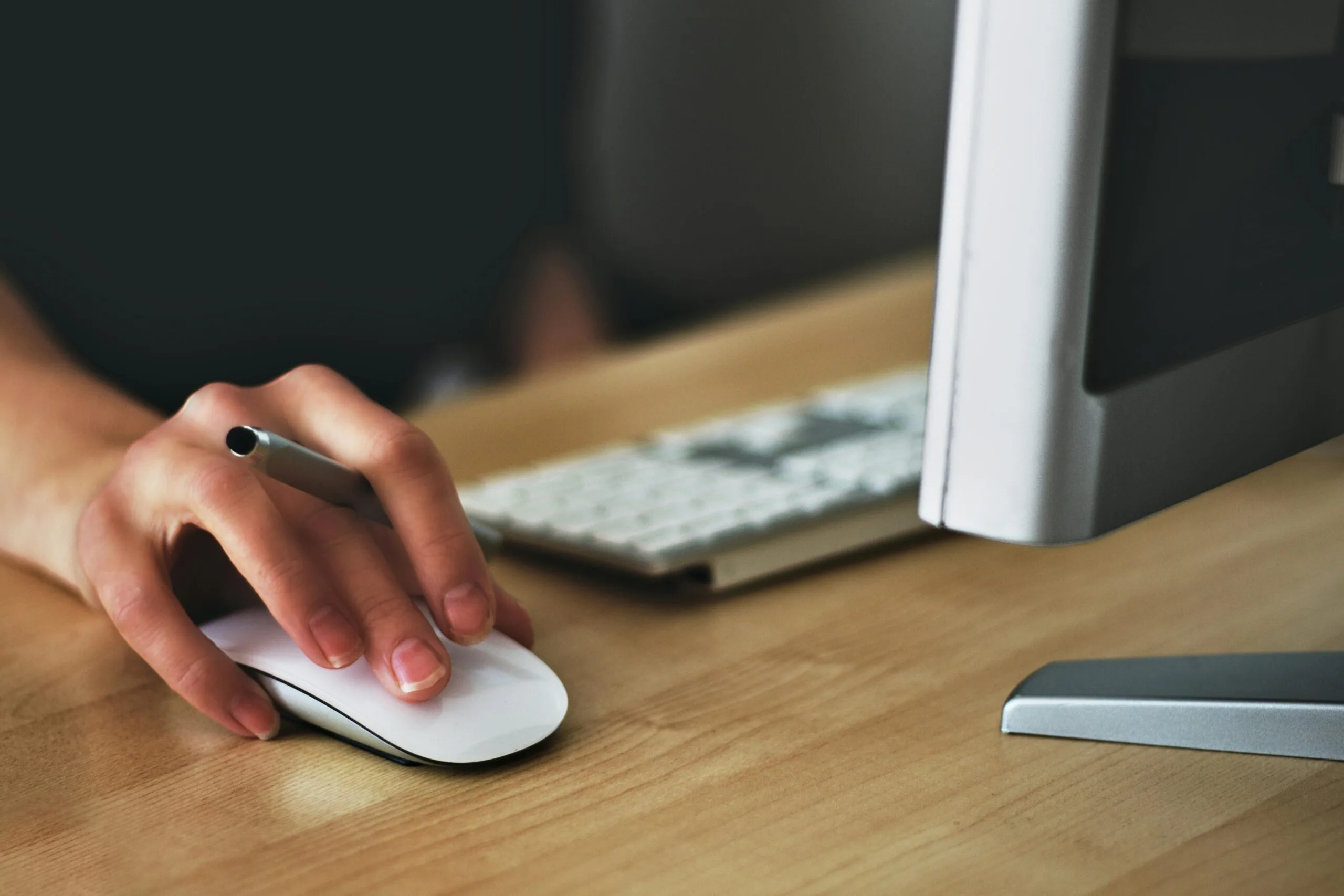 A hand holding a computer mouse while a keyboard and monitor are visible in the background.